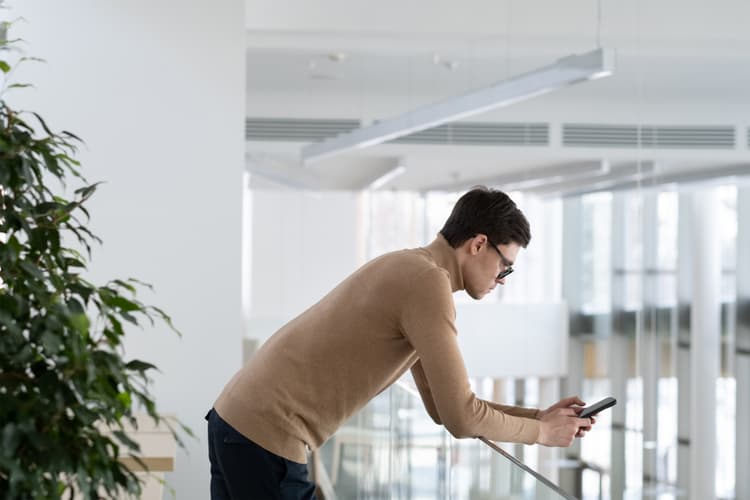 Person leaning over a railing while looking at a smartphone inside an office building.