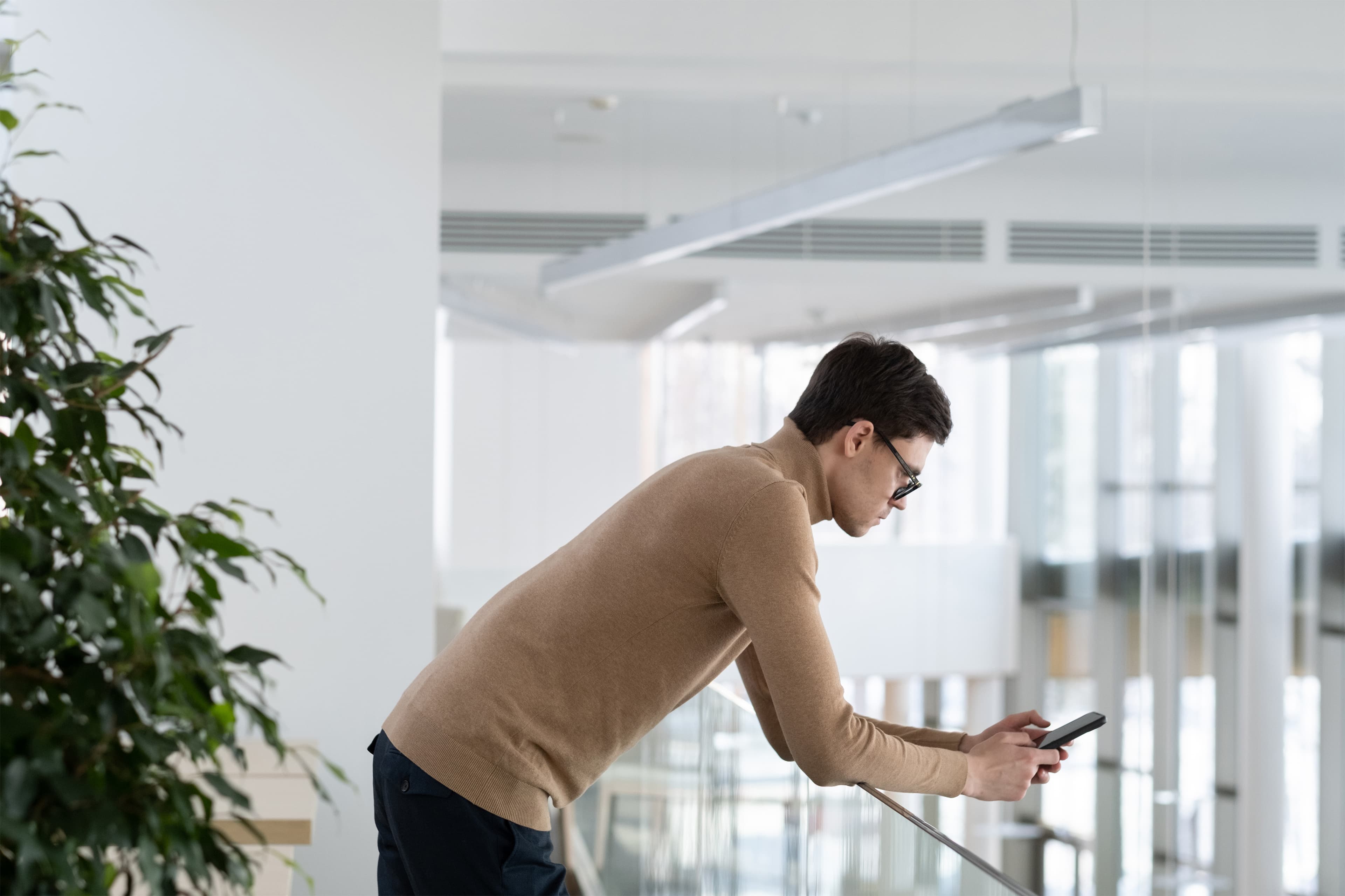 Person leaning over a railing while looking at a smartphone inside an office building.